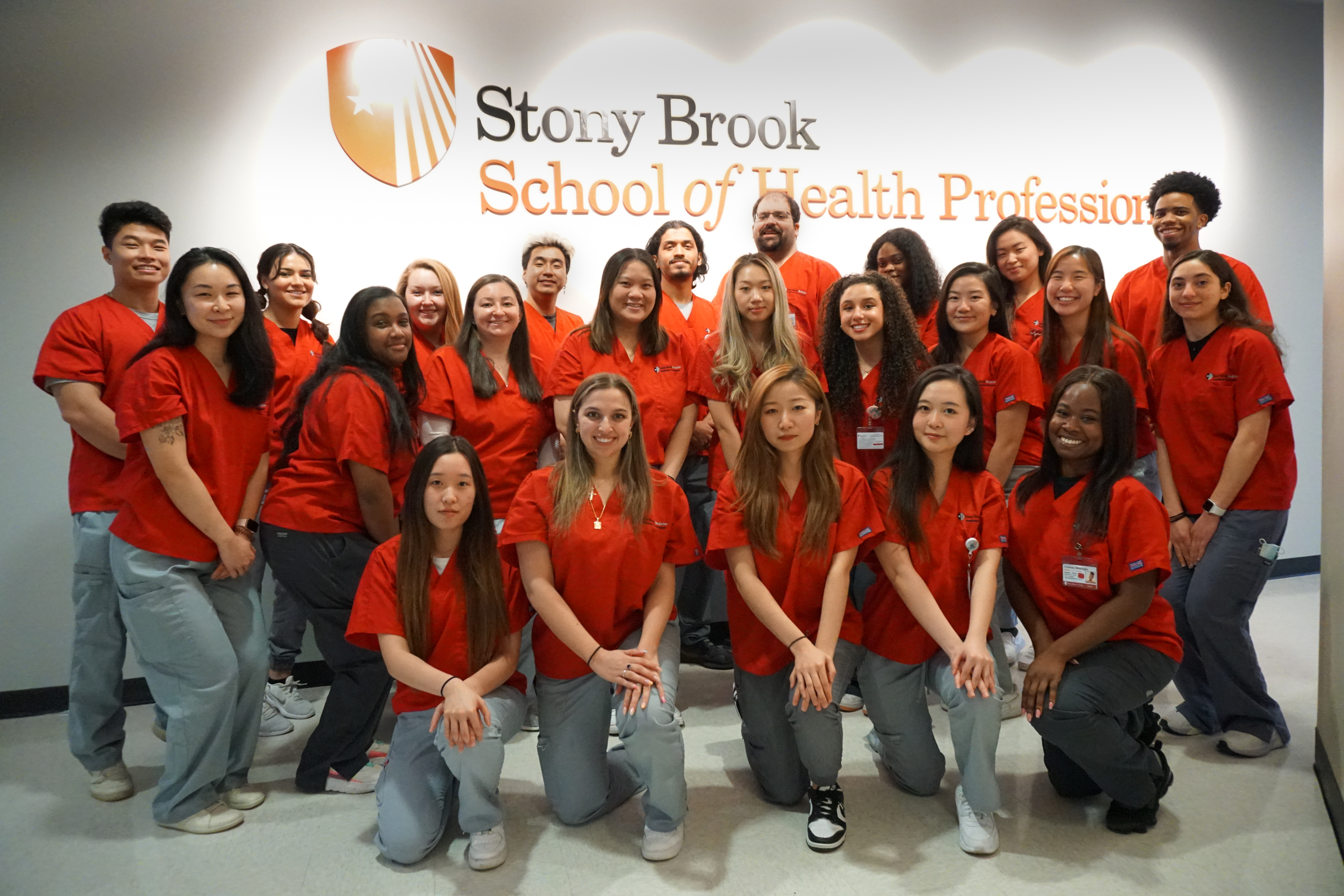 Photo of students in red shirt uniforms posing together in front of School of Health Professions logo sign