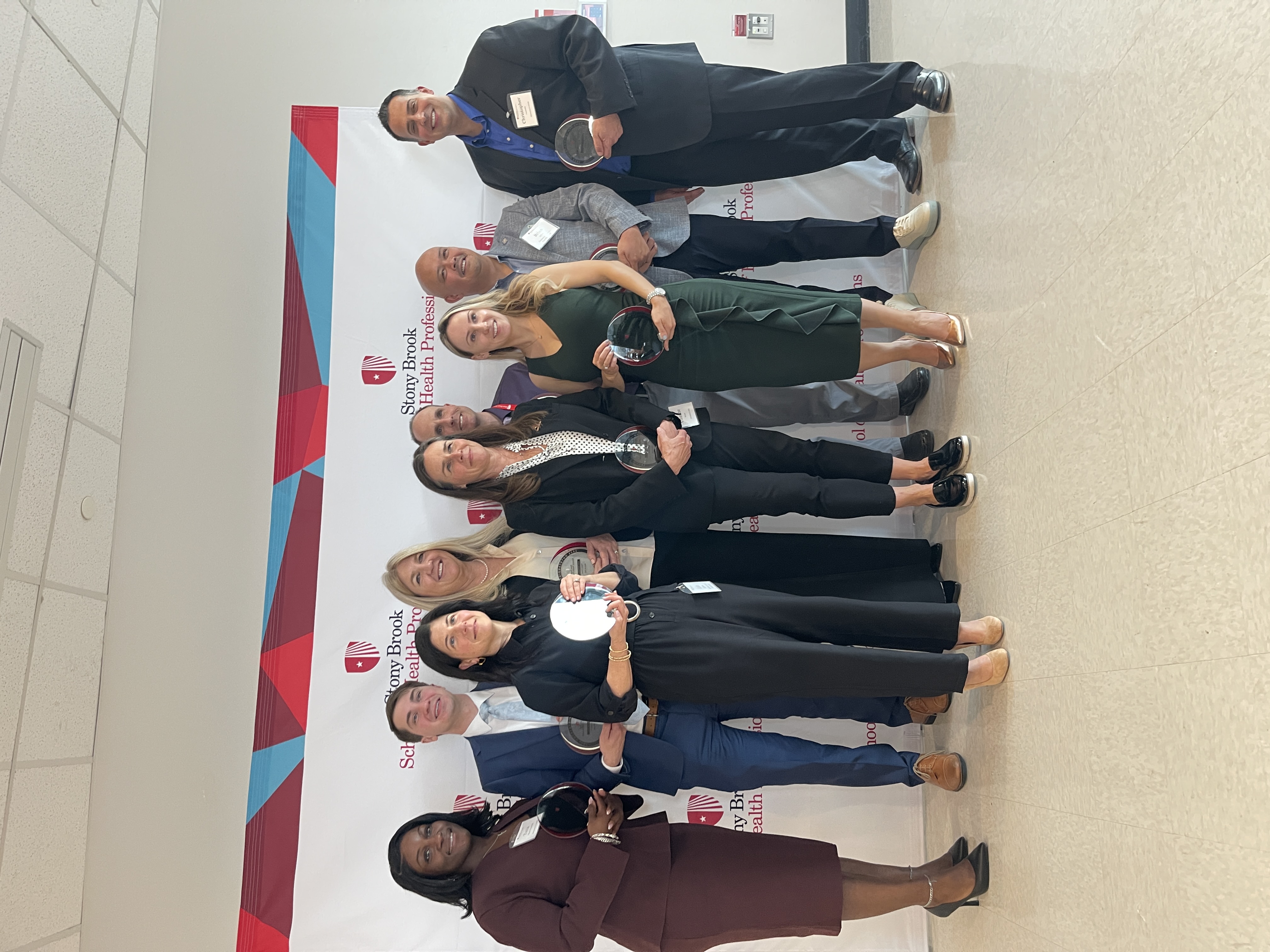 Nine smiling award recipients holding glass plaques and posing together in front of a Stony Brook School of Health Professions backdrop.