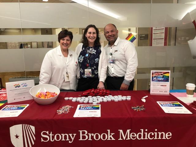 A group (Rose Cardin, Center: Dr. Allison Eliscu, Right: Dr. Adam Gonzalez)of people standing behind a table with food on it